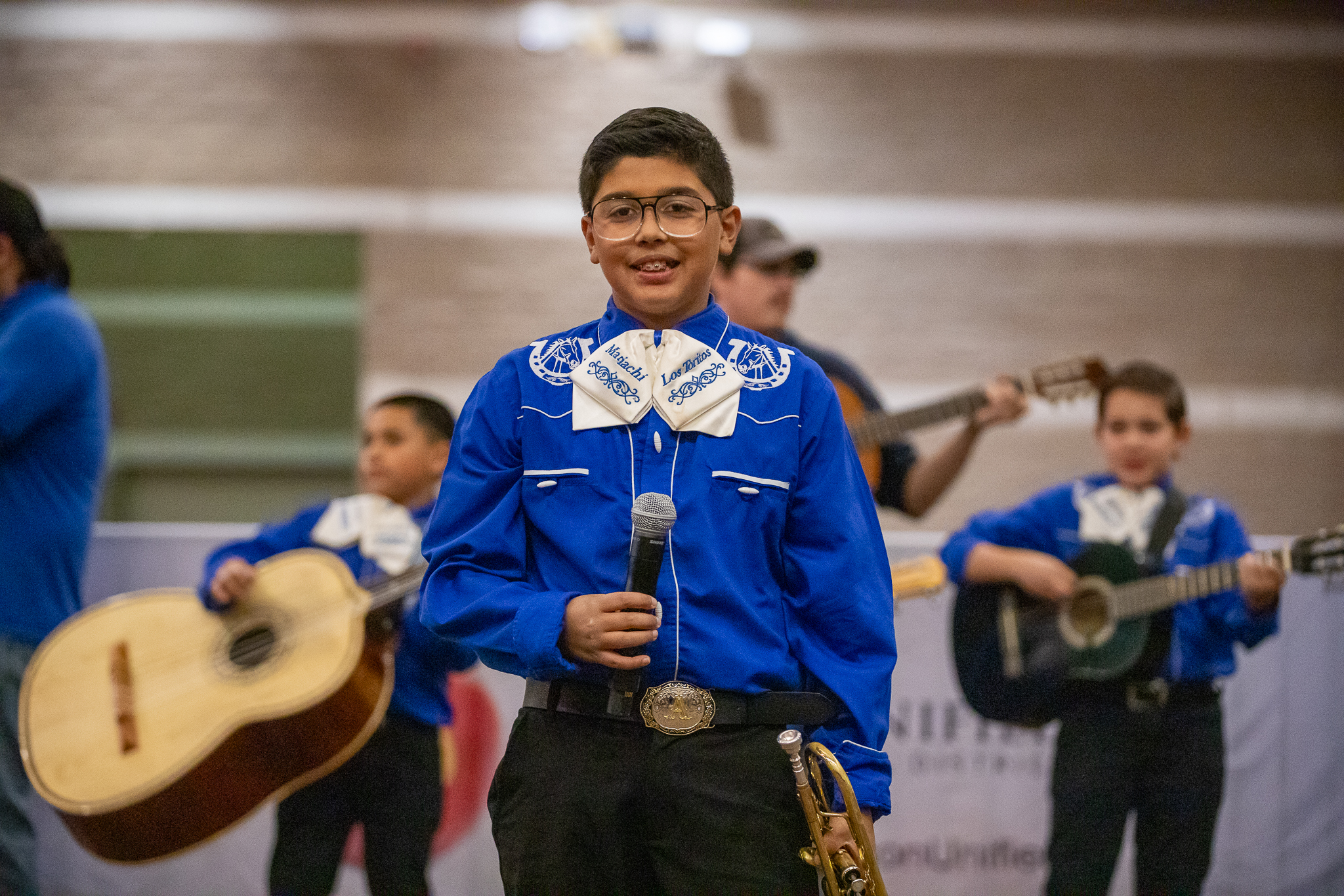 A boy in a blue dress shirt and glasses smiles with a microphone in his hand