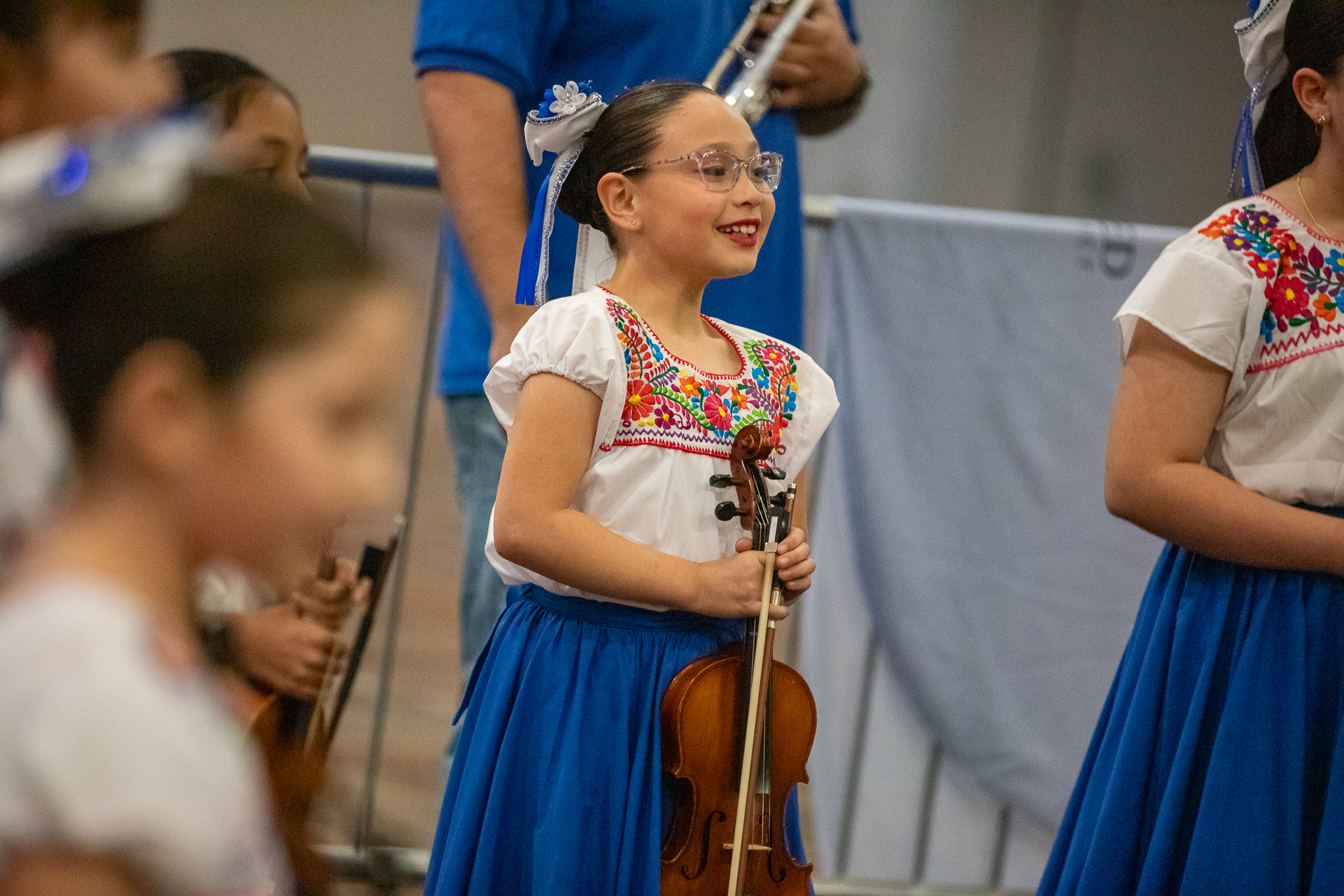 A girl in glasses and a blue and white folklorico dress smiles while holding her violin in front of her