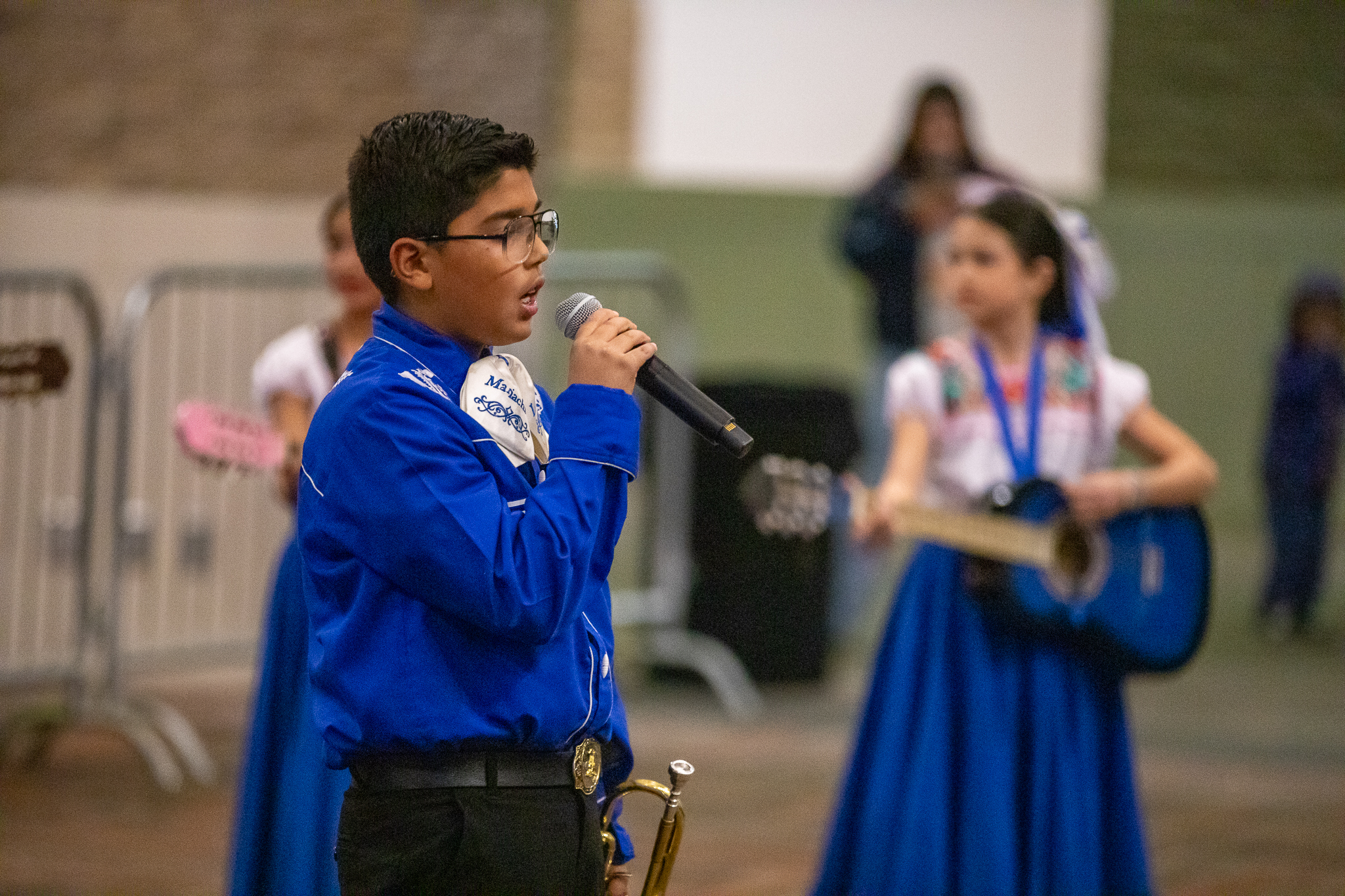 A boy in glasses and a blue dress shirt sings into a microphone