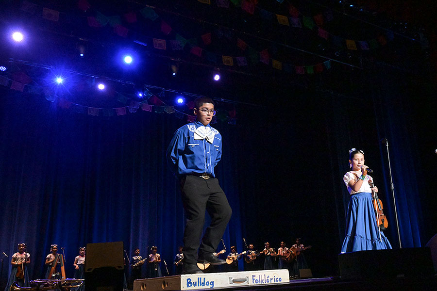 A boy dances at the front of the stage while a girl holding a violin sings