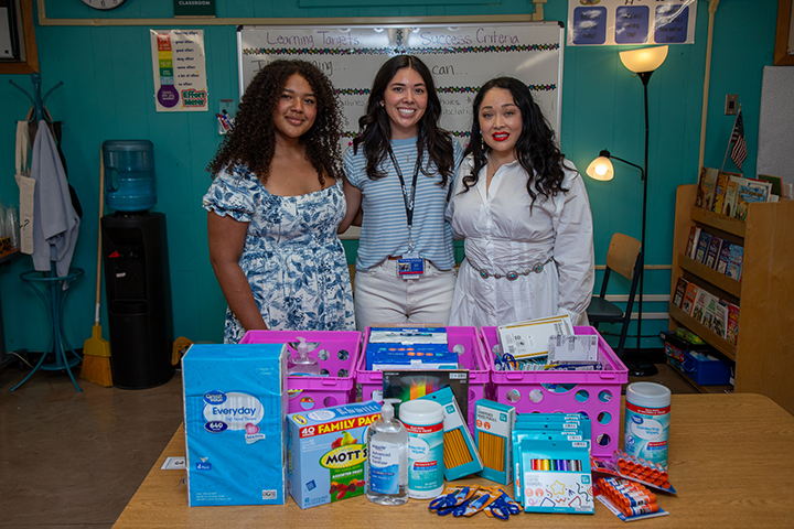 Three women smile behind a table filled with donated school supplies